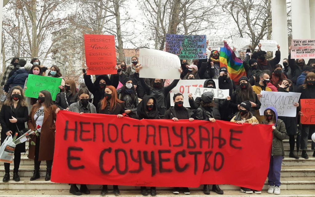 Masked protesters hold a red banner that says "Failure to Act Makes You Complicit" in Macedonian Cyrillic letters.