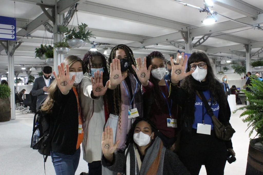A diverse group of young women wearing PP2 masks hold up their hands, on which are drawn eyes.