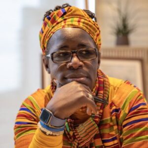 Rosamund Kissi Debrah sits in glasses and a colorful shirt and headwrap