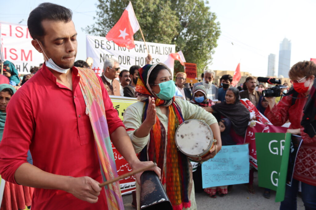 A man dressed in red bangs a cowbell at the front of a group of protesters at a demo in Karachi