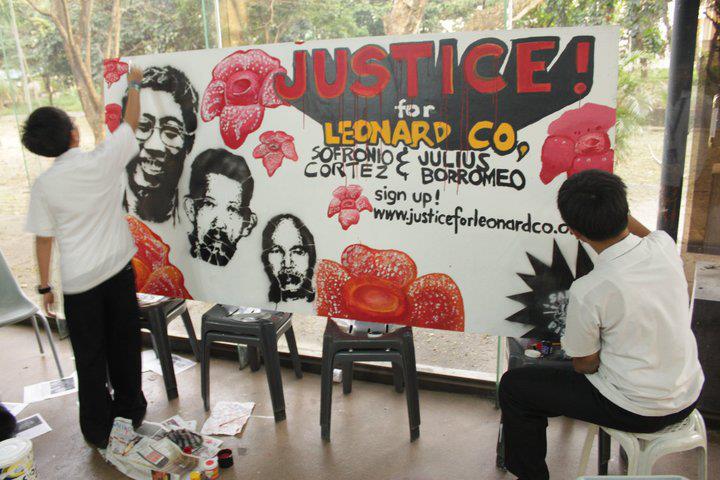 Artists stand around a large sign with pictures and flowers that reads "Justice for Leonard Co."