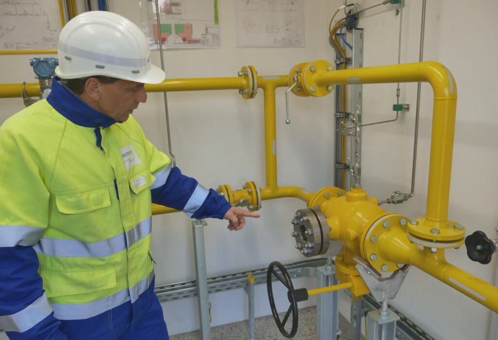 A man in a hardhat and jumpsuit points to yellow pipes conjoining in a hydrogen plant