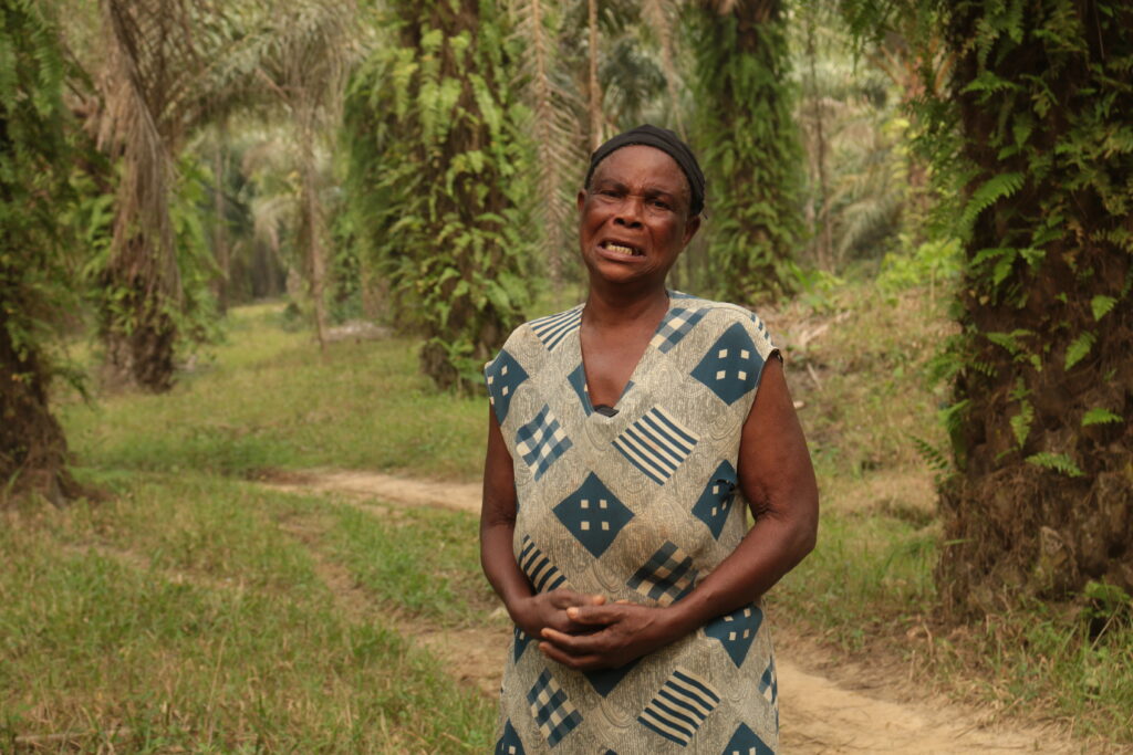 A woman stands in front of a lush plantation, she grimaces with her hands clasped in front of her waste.