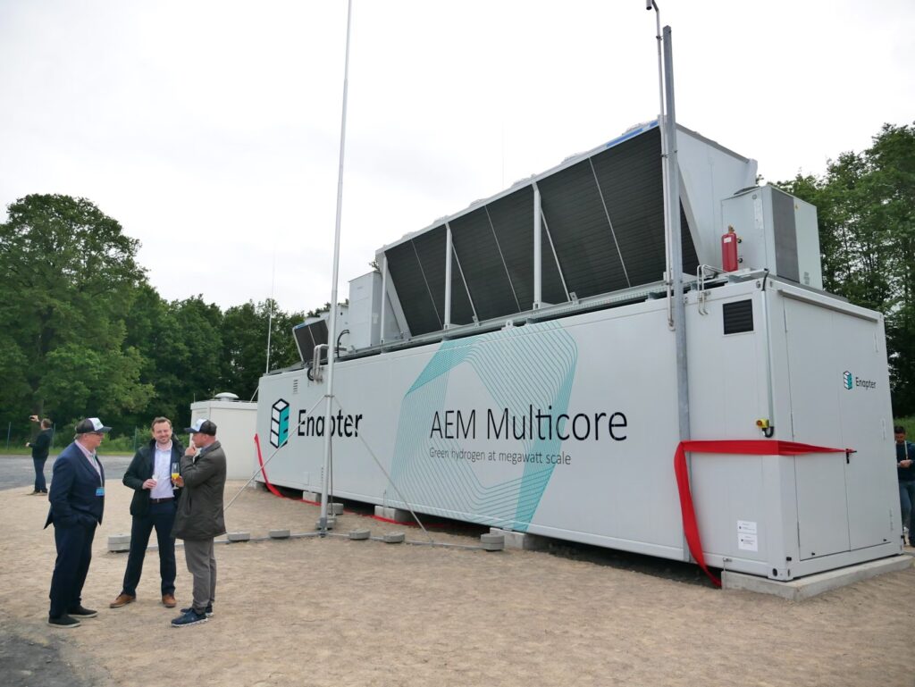 Men stand in front of a large metal box with grated panels towards the top, an electrolyzer, in Germany