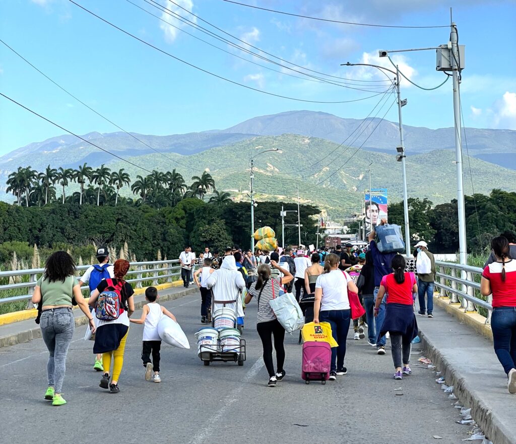 A group of people, mainly women, walk over a bridge with their belongings in suitcases and backpacks.