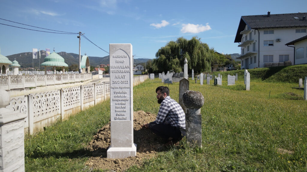 A man young kneels beside a grave column in a cemetary.
