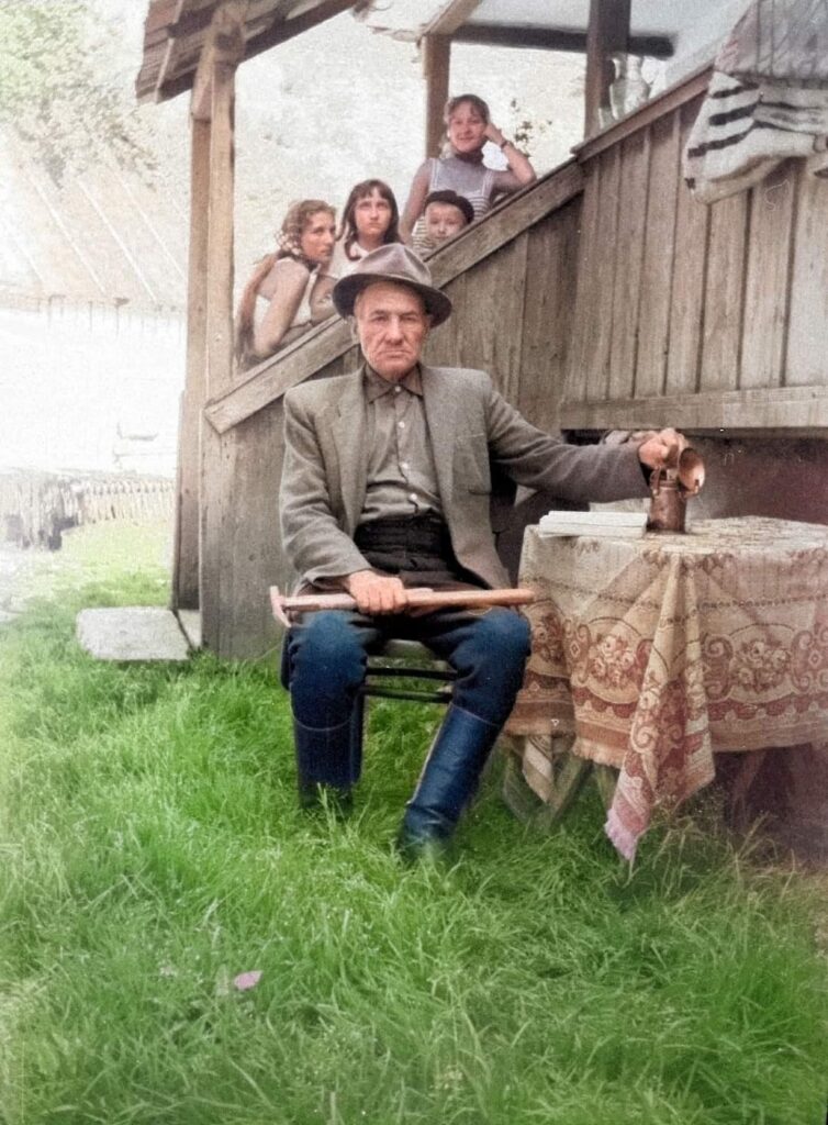 A older man sits in his backyard with a pickax, while granchildren peak over the railing behind him.