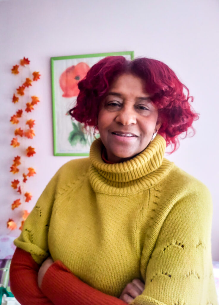 A woman with red hair and a yellow sweater looks at the camera confidently
