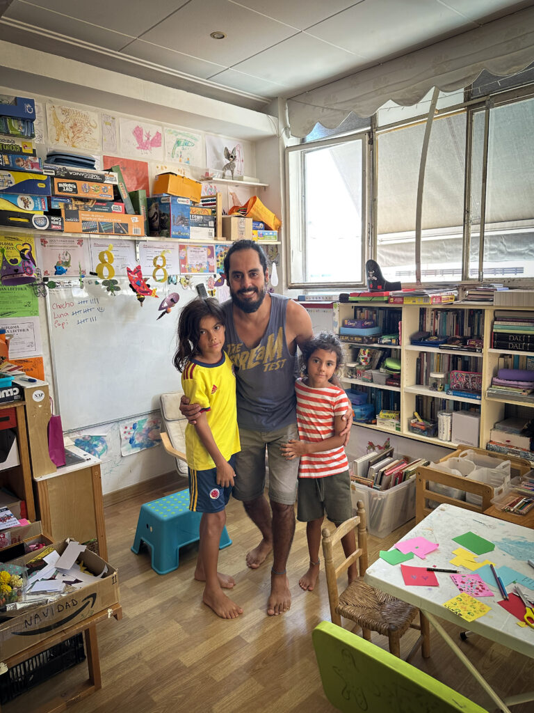 A man stand with his two children in a playroom in their apartment with shelves packed with toys and learning materials.