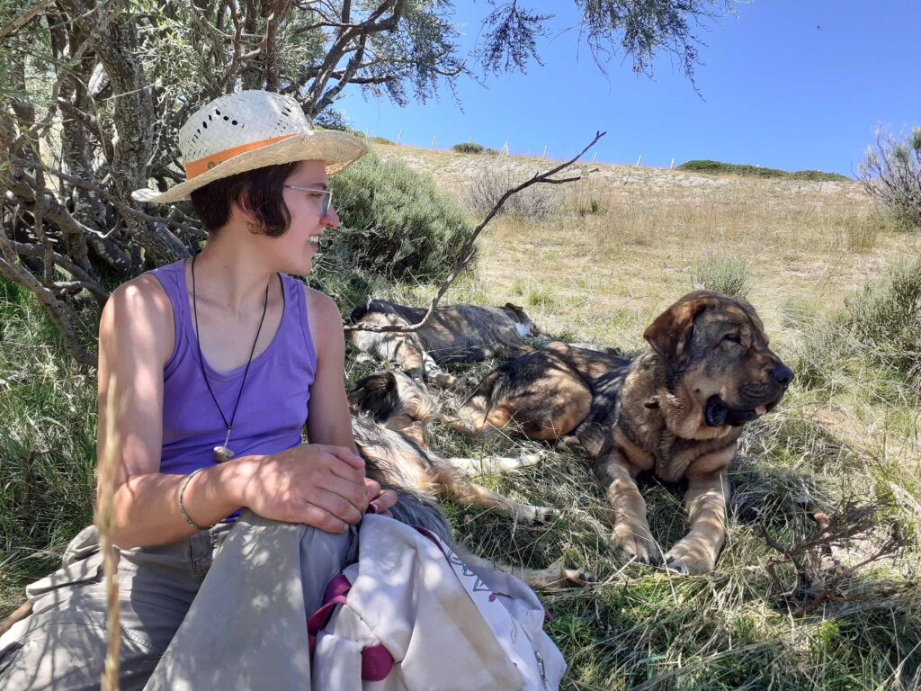 A young woman- Zuri- sits in a shady pasture next to a dog.